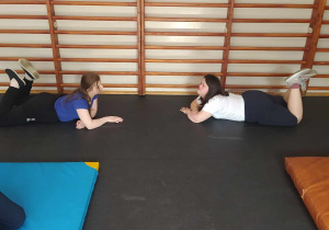 Na zdjęciu widać dwie osoby wykonujące ćwiczenia na macie gimnastycznej w sali gimnastycznej z drewnianymi drążkami na ścianie w tle. Osoby po lewej i po prawej stronie leżą na brzuchu z uniesionymi i zgiętymi w kolanach nogami.