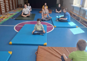 Grupa dzieci siedzących na matach w sali gimnastycznej. Ułożone są w pozycji lotosu, tradycyjnej pozycji jogi, która sprzyja równowadze i głębokiemu oddychaniu, których celem jest nauczenie dzieci uważności i prawidłowych technik oddychania.