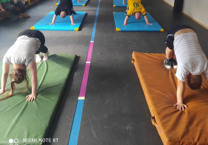 Grupa uczniów wykonujących ćwiczenia fizyczne podczas zajęć jogi (pozycja pieska). Ułożone są na kolorowych matach i pochylone do przodu, z rękami opartymi na ziemi. Zdjęcie wykonane w sali gimnastycznej z dużą ilością naturalnego światła, co wskazuje na spokojne i skupione miejsce na aktywność.