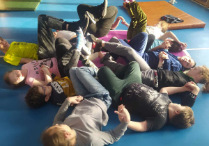 Grupa dzieci uczestniczących w zajęciach grupowych w sali gimnastycznej podczas zajęć relaksacyjnych. Dzieci leżą w kole na podłodze, z głowami na zewnątrz, tworząc z ciał okrąg, a nogi wyciągnięte do góry.