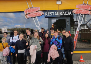 Na zdjęciu widzimy grupę osób stojących przed restauracją o nazwie “Porto-Grill.” Fasada restauracji jest żółta z niebieskimi akcentami, a po obu stronach szyldu znajdują się obrazy kebabów.